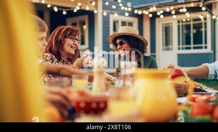 Aufgeregte multiethnische Frau, die mit einer Seniorin auf einer Sommergartenparty plaudert. Glückliche Gruppe von Kindern, Erwachsenen und älteren Menschen versammelte sich an einem Tisch, um Spaß zu haben und das Essen zu genießen. Stockfoto