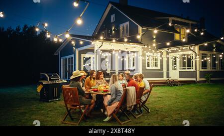 Eltern, Kinder und Freunde versammelten sich an einem Barbecue Dinner Tisch vor einem wunderschönen Haus mit Lichtern Dekorationen. Alte und junge Leute haben Spaß und essen Essen. Gartenfeier im Garten. Stockfoto