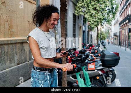 Ein junger Mann mietet einen Roller über eine mobile Anwendung. Ein gutaussehender, zufriedener Kerl nutzt umweltfreundliche städtische Verkehrsmittel, mietet einen Roller mit seinem Handy Stockfoto