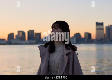 Japanische Frau im Mantel, die am Wasser steht, mit einer Stadt im Hintergrund Stockfoto