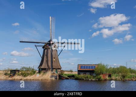 Windmühle neben einem kleinen Haus auf einem Wasserlauf unter einem blauen Himmel mit weißen Wolken, historische Windmühlen auf einem Fluss vor einem blauen Himmel, Kinderdijk Stockfoto