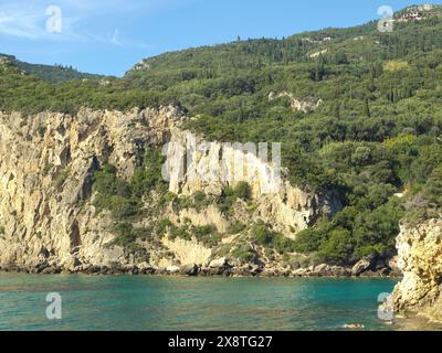 Steile Klippen und bewaldete Hügel entlang der felsigen Küste unter blauem Himmel, kleiner Kieselstrand zwischen felsigen Bergen mit Sonnenschirmen und Booten, Korfu Stockfoto