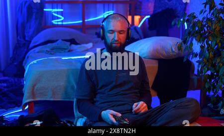 Junger bärtiger blonder Mann, der auf dem Boden neben seinem Bett sitzt. Medien. Mann schüttelt den Kopf, während er Musik im Schlafzimmer mit Neonlampen hört. Stockfoto