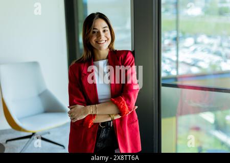 Die junge Frau steht selbstbewusst mit einem Lächeln in einem stilvollen roten Blazer und lehnt sich in einem modernen Bürobereich an ein Glasfenster Stockfoto