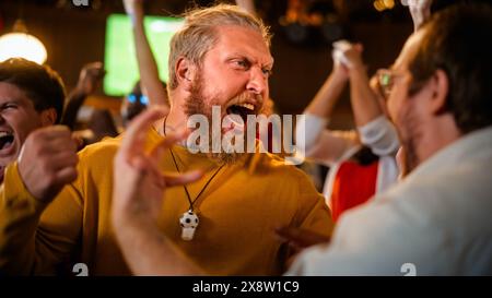 Begeisterte Soccer Club-Mitglieder jubeln ihr Team in einem Pub an. Unterstützende Fans, die in einer Bar stehen, jubeln, die Hände heben und schreien. Freunde feiern den Sieg nach dem Tor. Stockfoto