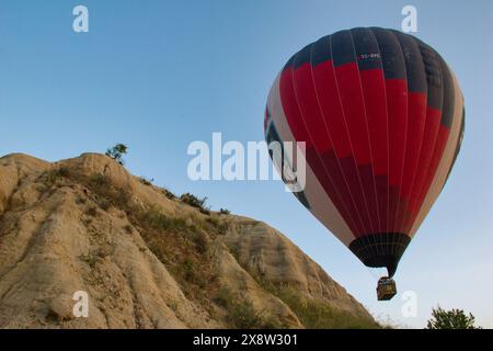 Ein beeindruckender Heißluftballon gleitet über das zerklüftete Gelände von Kappadokien, Türkei, unter einem hellblauen Himmel. Das Bild fängt das Wesen des Abenteuers ein Stockfoto