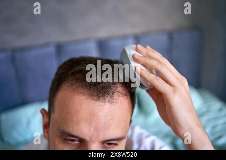 Der Mann massiert seinen Kopf mit einem elektrischen Massagegerät für Haarwuchs Stockfoto