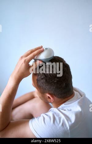 Der Mann massiert seinen Kopf mit einem elektrischen Massagegerät für Haarwuchs Stockfoto