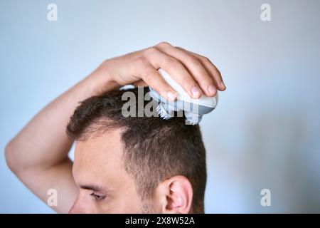 Der Mann massiert seinen Kopf mit einem elektrischen Massagegerät für Haarwuchs Stockfoto