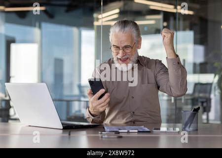 Ein älterer Mann mit grauem Haar feiert Erfolg in einem modernen Büro und blickt freudig auf sein Smartphone Stockfoto
