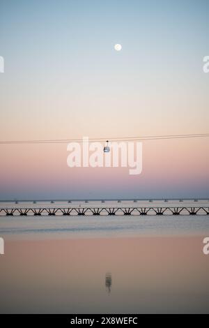 Ein vertikaler Panoramablick auf eine Seilbahn, die über ein ruhiges Gewässer mit einer Brücke im Hintergrund fährt, während eines ruhigen Sonnenuntergangs. Die SKY tr Stockfoto