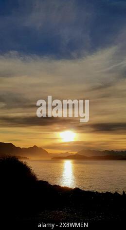 Atemberaubender Sonnenuntergang über einem ruhigen Meer mit der Reflexion der Sonne auf dem Wasser und den Bergen in der Ferne unter einem bewölkten Himmel Stockfoto