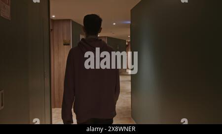 Junger Mann in rosa Oversize-Hoodie im Hotel. Stock Clip. Ein Mann, der an Zimmertüren entlang eines langen Korridors läuft. Stockfoto