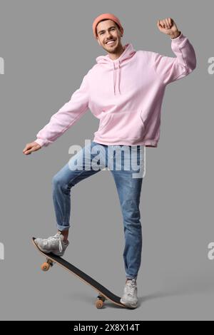 Hübscher, lächelnder junger Mann in stilvollem rosa Hoodie und pfirsichfarbenem Hut mit Skateboard, der auf grauem Hintergrund posiert Stockfoto