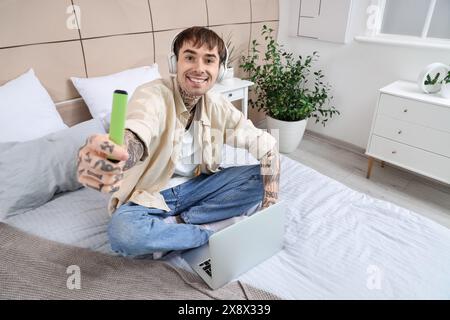 Junger gutaussehender Mann in Kopfhörern mit elektronischer Wegwerfzigarette, der Laptop auf dem Bett zu Hause benutzt Stockfoto