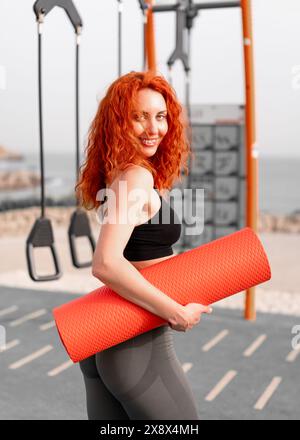 Lächelnde rothaarige Sportlerin mit gerollter Yogamatte. Seitenansicht der Passform starke Sportlerin in Sportbekleidung mit Blick auf die Kamera beim Stehen im Fitnessstudio im Freien an sonnigen Tagen. Stockfoto