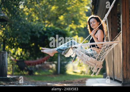 Glückliche junge Frau, die auf einem Makrame-Schaukelstuhl in der Nähe des Landhauses im Freien sitzt Stockfoto