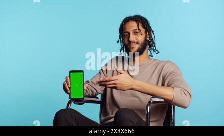 Mann aus dem Nahen Osten im Rollstuhl, der auf das Mobiltelefon mit grünem Bildschirm zeigt. Person mit Querschnittslähmung mit Chroma-Schlüssel-Handy, isoliert über blauem Studiohintergrund Stockfoto