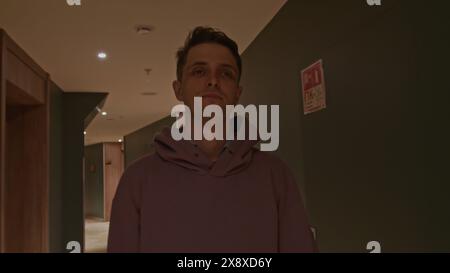 Junger Mann in rosa Oversize-Hoodie im Hotel. Stock Clip. Ein Mann, der an Zimmertüren entlang eines langen Korridors läuft. Stockfoto