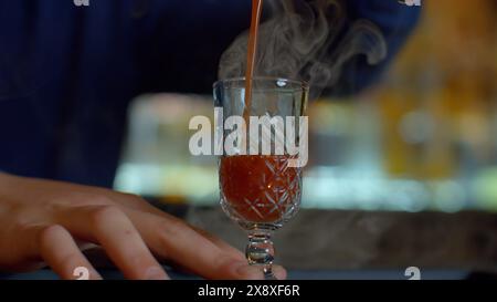 Nahaufnahme eines Barkepers an einer Bar, der Alkohol in ein Vintage-Glas gießt. Medien. Gießen Sie rote heiße alkoholische Tinktur mit Dampf. Stockfoto