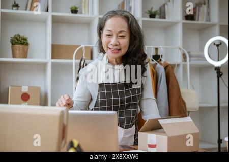 Eine attraktive reife asiatische weibliche Online-Bekleidungsläuferin, die Online-Bestellungen auf ihrem Computer überprüft und in ihrem Heimbüro arbeitet. E-Commerce, Mode s Stockfoto