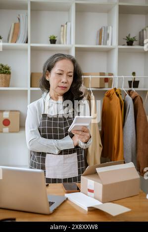 Eine gestresste und ernsthafte, reife asiatische Online-Verkäuferin berechnet ihre Ausgaben für Marketinganzeigen auf ihrem Rechner, während sie in ihrem Heimbüro arbeitet. Stockfoto