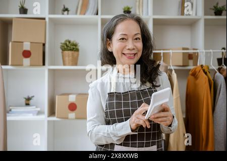 Eine attraktive, reife asiatische Verkäuferin im Online-Shop hält einen Taschenrechner und lächelt in die Kamera, während sie in ihrem Heimbüro steht. Elektronischer Geschäftsverkehr, Stockfoto