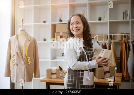 Eine attraktive, positive, reife asiatische Online-Verkäuferin, die Kartons hält und von der Kamera weglächelt, während sie in ihrem Büro steht. Onlin Stockfoto