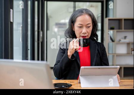 Eine selbstbewusste, reife asiatische Geschäftsfrau ist darauf konzentriert, Online-Dokumente auf ihrem digitalen Tablet zu lesen, während sie am Schreibtisch im Büro Kaffee trinkt. Stockfoto