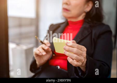 Ein selektives Bild einer nachdenklichen asiatischen Geschäftsfrau, die eine Haftnotiz und einen Stift in der Hand hält, die an ihrem Schreibtisch im Büro in Gedanken verloren ist. Stockfoto