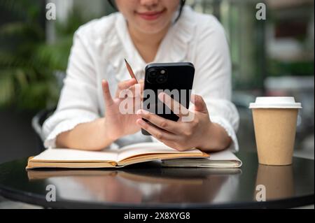 Ein zugeschnittenes Bild einer jungen positiven asiatischen Frau, die ihr Smartphone benutzt, während sie an einem Tisch etwas in ihr Notebook schreibt. Hausaufgaben machen, zu tun machen Stockfoto