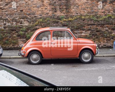 Cremona, Italien - 15. Mai 2024 der elegante, altmodische orangene Fiat 600 Wagen aus dem italienischen Jahr 60 hebt sich vor einer rustikalen Ziegelmauer hervor und erinnert an Nostalgie an einem Kai Stockfoto