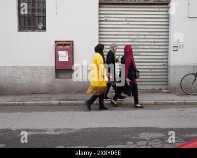 Cremona, Italien - 15. Mai 2024 drei muslimische Frauen in traditioneller Kleidung gehen an einer geschlossenen Ladenfront mit Metallverschluss vorbei und zeigen alltägliche c Stockfoto