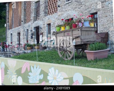 Morfasso , Italien - 15. Mai 2024 malerischer Vintage-Holzwagen mit bunten Blumen vor einem alten Steingebäude mit charmanten Holzdetai Stockfoto