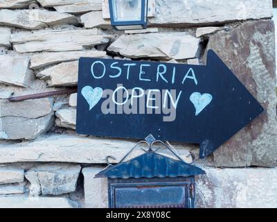 Einladendes schwarzes Pfeilschild mit weißer Schrift, das eine offene Osteria anzeigt, montiert auf einem rustikalen Steinmauerhintergrund Stockfoto