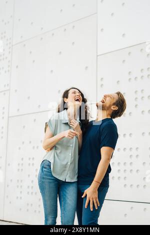 Ein Paar lacht zusammen, während es vor einer modernen weißen Wand mit kreisförmigen Mustern steht. Sie genießen eindeutig die Gesellschaft und das Teilen des anderen Stockfoto