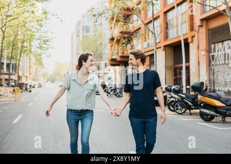 Ein Paar läuft Hand in Hand die Straße der Stadt entlang, lächelt und schaut sich an und genießt einen sonnigen Tag. Stockfoto