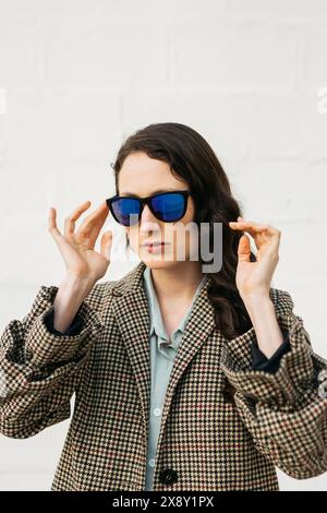 Porträt einer Frau, die ihre Sonnenbrille anpasst, während sie einen Hahnentrittmantel trägt und an einer weißen Wand steht. Stockfoto