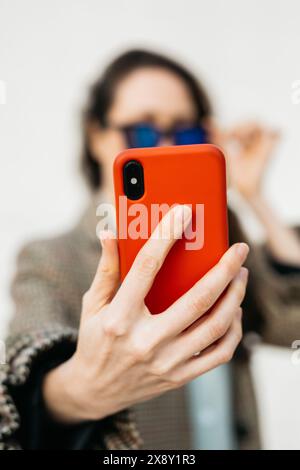 Frau mit Sonnenbrille und kariertem Mantel, die ein Selfie mit ihrem roten Smartphone macht und ihre Brille mit einer Hand anpasst. Stockfoto