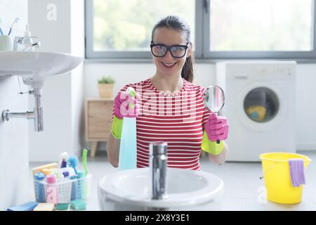 Glückliche Frau, die von Hygiene besessen ist: Sie hält ein Waschmittel und kontrolliert Oberflächen mit einer Lupe Stockfoto