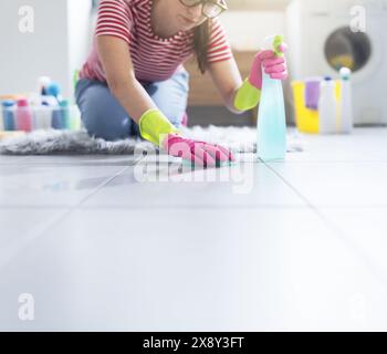 Frau, die zu Hause Hausaufgaben macht, kniet auf dem Boden und reinigt die Fliesen mit einem Schwamm Stockfoto