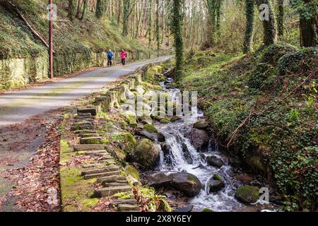 Stream läuft neben einer Landstraße mit zwei Frauen, die hinunter laufen. Bangor, Gwynedd, Nordwales, Großbritannien Stockfoto