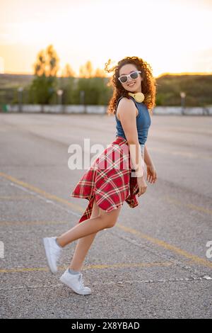 Fröhliche junge Frau in Sonnenbrille und Kopfhörern, die bei Sonnenuntergang auf einem Parkplatz spielerisch herumwirbelt. Stockfoto