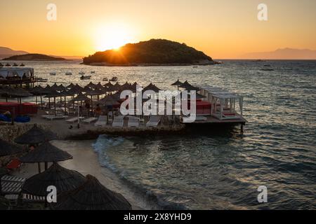 Romantische Landschaft des Piers mit Sonnenliegen, dem Ionischen Meer und der Insel Ksamil während der Goldenen Stunde an der albanischen Riviera. Strandküste in Albanien bei Sonnenuntergang. Stockfoto
