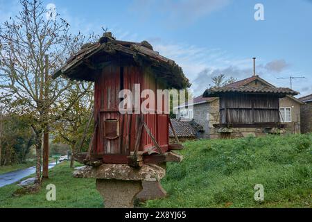 Das alte, rote hölzerne horreo steht auf einem Steinfuß in einer ländlichen Landschaft mit bewölktem Himmel und Vegetation im Hintergrund Stockfoto