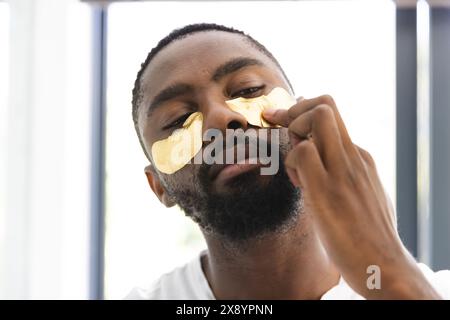 Zu Hause, ein Afroamerikaner, der Gold unter den Augen aufträgt. Mit kurzen schwarzen Haaren und Bart, mit weißem Hemd und schwarzem Hintergrund Stockfoto
