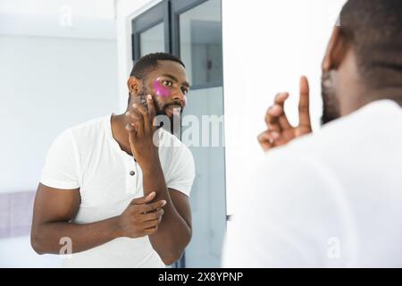 Zu Hause, ein Afroamerikaner, der Gesichtsmaske aufträgt und in einen Spiegel blickt Stockfoto