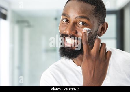 Zu Hause, Afroamerikaner, der Feuchtigkeitscreme aufträgt, lächelt in die Kamera. Mit Bart und kurzen Haaren trägt er ein weißes Hemd und strahlt entspannt aus Stockfoto