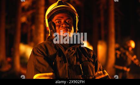 Porträt eines tapferen gutaussehenden jungen erwachsenen Feuerwehrmannes in Sicherheitsuniform und einem Helm, der während eines Wildfeuers für die Kamera posiert. Professioneller schwarzer Feuerwehrmann, der die Kamera ansieht und lächelt. Stockfoto