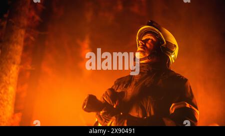 Ein erfahrener Feuerwehrmann löscht ein Wildlandfeuer tief in den Wäldern. Profi in Sicherheitsuniform und Helm, der den Feuerschlauch hält, nach oben blickt, die Windrichtung annimmt und berechnet. Stockfoto